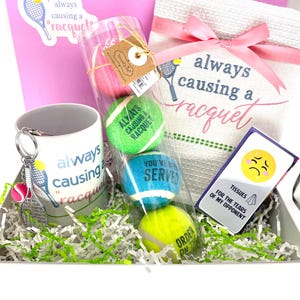 May include: A tennis-themed gift set in a white box, featuring a mug, keychain, tennis balls, and a towel. The mug and towel have the phrase "always causing a racquet." The set includes a card that says "Tissues for the tears of my opponent."