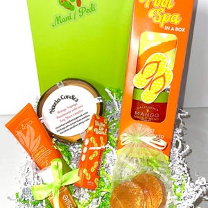 May include: A gift box with a green background and various mango-scented spa products. Includes a shampoo, lip balm, candles, and a foot spa kit with a towel and flip-flop design. The box is filled with green paper.