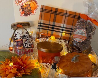 May include: A gift box filled with fall-themed items. Includes a jar with a coffee design, a plaid orange and black blanket, a pumpkin-shaped mug, a candle, and chocolate-covered pretzels. The box is decorated with fairy lights and a large orange flower.