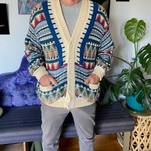 May include: A cream-colored cardigan with a blue, red, and green geometric pattern. The cardigan has a V-neck and a button closure.