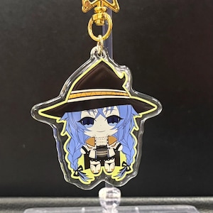 May include: A clear acrylic keychain featuring a cartoon witch with blue hair and a black hat with yellow trim. The keychain has a gold-colored star-shaped clasp and a gold-colored ring.