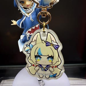 May include: A clear acrylic keychain featuring a cartoon character with blonde hair, blue eyes, and a sailor-style outfit. The keychain is attached to a gold-colored metal clasp. A figurine of a character in a blue shark hoodie is in the background.