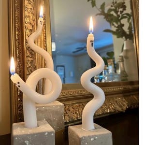May include: White ghost-shaped candles with black dot eyes and mouths, atop gray square concrete bases. The candles have a whimsical, wavy design and are lit with a warm flame. The background includes a gold-framed mirror.