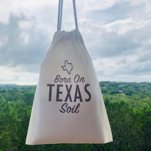May include: A white drawstring bag with the text "Born On Texas Soil" and an outline of the state of Texas. The bag is hanging from a white string against a cloudy sky background.