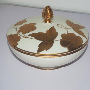 Peut inclure: Un bol en céramique blanc avec un couvercle orné de feuilles et de motifs floraux dorés. Le couvercle est surmonté d'un bouton doré. Le bord et la base du bol sont également dorés. Une pièce décorative pour servir ou exposer.