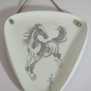 Op de afbeelding: Witte, driehoekige decoratieve plaat met een grijze en zwarte inkttekening van een steigerend paard. De plaat heeft een goudkleurig touw om op te hangen. De paardenillustratie is in een traditionele Aziatische kunststijl.