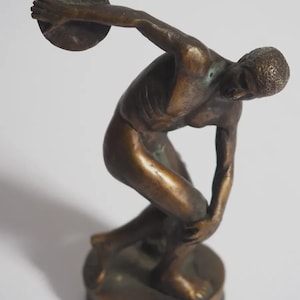 Könnte beinhalten: Bronzeskulptur eines Diskuswerfers in Bewegung. Die Figur ist mit muskulösem Körperbau dargestellt und bereit, den Diskus zu werfen. Die Skulptur hat eine dunkle, gealterte Patina und ist auf einem kreisförmigen Sockel montiert. Die Skulptur ist etwa 15 cm hoch.