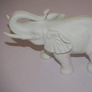May include: A white ceramic elephant ornament with its trunk raised. The elephant has detailed features, including large ears and tusks. The ornament is a decorative item, suitable for a home or office.