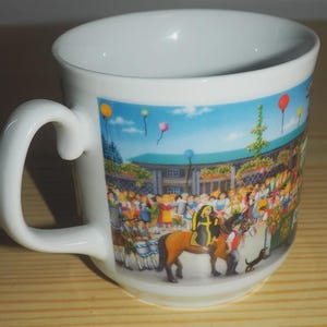 Może przedstawiać: Biały ceramiczny kubek z zakrzywionym uchwytem. Kubek przedstawia kolorową ilustrację tłumu ludzi, konia i balonów na tle błękitnego nieba. Scena wydaje się być festiwalem lub paradą.