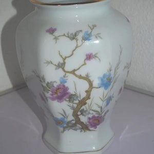 Peut inclure: Vase en céramique blanche avec un bord doré et une base hexagonale. Le vase est décoré d'un motif floral avec des fleurs violettes et bleues, et une branche marron. C'est un objet décoratif.