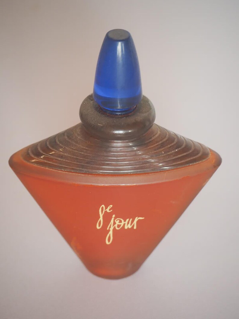 Op de afbeelding: Een vintage parfumflesje met een unieke vorm. De fles is warm oranje van kleur met een blauwe dop. De woorden "de jour" staan in gouden letters op de voorkant. De fles heeft een gelaagd ontwerp.