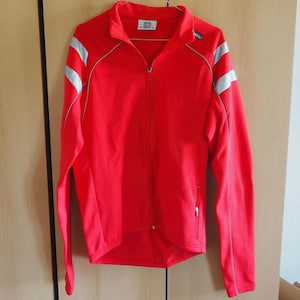 Peut inclure: Une veste de sport rouge vif à manches longues avec une fermeture éclair intégrale, un col montant et deux bandes réfléchissantes argentées sur chaque manche. Le vêtement présente une petite étiquette blanche au col et un logo sur la poitrine.