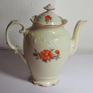 Puede incluir: Una tetera de porcelana color crema con borde festoneado y tapa a juego. El cuerpo y la tapa están decorados con motivos florales de rosas naranjas y hojas verdes. Los bordes de la tapa, el pico, el asa y la base tienen finas líneas naranjas.