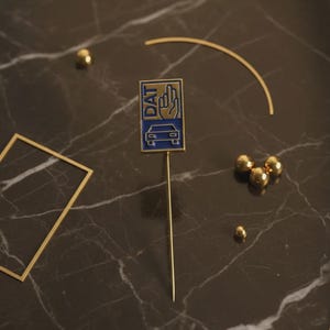 Può includere: Spilla dorata con un emblema rettangolare blu e oro con la scritta "DAT", un gesto della mano e una silhouette di auto. La spilla è su un lungo e sottile stelo dorato. Sono presenti anche sfere dorate e una cornice quadrata.