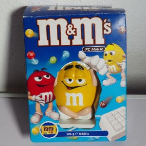 以下が含まれることがあります： 中央に黄色いM&Mキャラクター型のマウスが入った、M&M's PCマウス用の青い段ボール箱。パッケージには左側に赤いM&Mキャラクター、右側に黄色いM&Mキャラクター、そして「M&M's PC Mouse 125 g」というテキストが印刷されています。