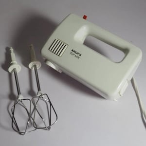 Op de afbeelding: Een witte Krups Top Mix handmixer met een rechthoekige behuizing, een handvat en een kleine rode schakelaar aan de bovenkant. Twee afneembare metalen gardes met draadlussen en witte kunststof schachten liggen naast de mixer op een ondergrond.