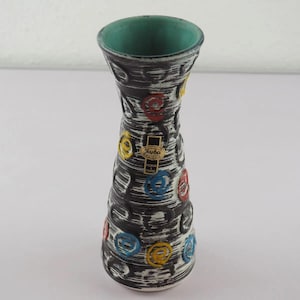 Può includere: Un vaso in ceramica alto con bordo svasato e interno color ottanio. L'esterno presenta una finitura testurizzata bianca e nera con motivi a spirale in rilievo sparsi in rosso, giallo e blu. È presente un'etichetta Jasba.