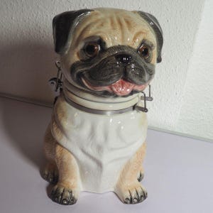 May include: A ceramic pug-shaped jar with a hinged lid and metal clasp. The jar is tan and black, with a white chest and a pink tongue. The pug is sitting upright, with its paws visible.