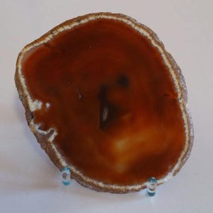 Pode incluir: Uma fatia de ágata polida, com um centro castanho-avermelhado profundo e uma borda externa mais clara, de cor creme. A pedra natural apresenta um padrão único e é exibida em um suporte transparente.