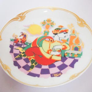 Pode incluir: Prato decorativo de cerâmica com uma ilustração colorida. O design apresenta uma figura com roupas vermelhas e amarelas, um chão xadrez e uma borda dourada. O prato tem um estilo caprichoso e artístico.