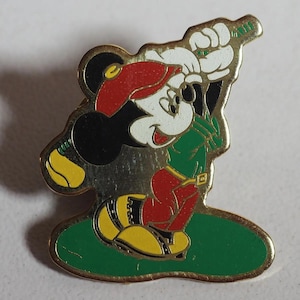 Könnte beinhalten: Goldfarbene Anstecknadel mit Mickey Mouse in Golfkleidung. Mickey trägt einen roten Hut, ein grünes Hemd, eine rote Hose und gelbe Schuhe. Die Anstecknadel befindet sich auf einem grünen Golfplatz, Mickey hält einen Golfschläger.