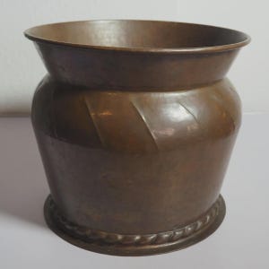 Puede incluir: Una maceta redonda de metal en tono cobre oscuro con un borde ancho y acampanado, y un cuerpo decorado con crestas diagonales en relieve. La base del recipiente está rodeada por un borde elevado con un diseño de cuerda trenzada.