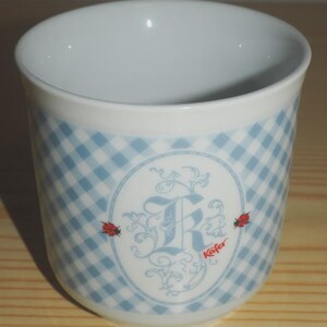 May include: A white ceramic cup with a light blue gingham pattern. The cup features an oval design with a stylized letter "R" and the word "Käfer". Two red ladybugs are on either side of the oval design.