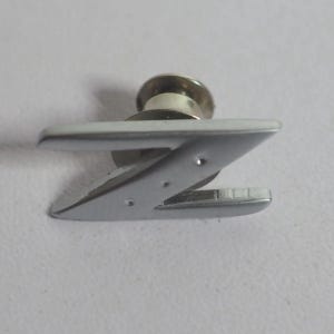 Könnte beinhalten: Eine silberfarbene Anstecknadel aus Metall in Form des Großbuchstabens Z. Die Nadel hat eine glatte, reflektierende Oberfläche mit metallischem Glanz und verfügt über einen runden Verschlussmechanismus auf der Rückseite zur sicheren Befestigung.