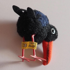 Könnte beinhalten: Ein Vintage-Steiff-Vogel-Plüschtier. Der Vogel hat ein schwarzes, strukturiertes Gefieder mit einem blauen Fleck auf dem Rücken. Er hat einen leuchtend orangefarbenen Schnabel und rote Beine und Füße. Ein gelbes Etikett mit Text ist an seinen Beinen befestigt.