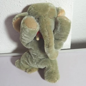 Könnte beinhalten: Ein Plüschelefant mit weichem, salbeigrünem Fell, großen Ohren mit beigen Innenseiten, einem nach oben gerichteten Rüssel und einem kleinen roten Band um den Hals. Das Stofftier hat eine strukturierte, samtige Oberfläche und Optik.