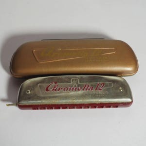 May include: A silver and red Chrometta 12 M. Hohner harmonica with a metallic finish, positioned in front of a matching gold-toned hard carrying case. Both items feature the embossed text Chrometta 12 and M. Hohner Made in Germany.