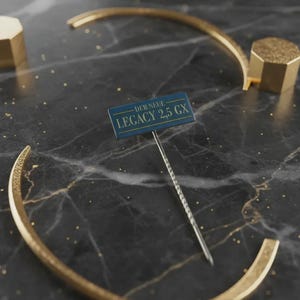 Può includere: Un oggetto metallico dorato curvo e due forme geometriche dorate sono disposti su una superficie di marmo nero con venature dorate. Un piccolo cartello con la scritta "DER NEUE LEGACY 2,5 GX" è al centro.