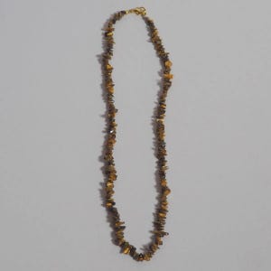 Peut inclure: Un collier composé de nombreuses petites pierres semi-précieuses en œil-de-tigre, de forme irrégulière et polie, dans des tons brun doré, ambre et gris anthracite. Les pierres sont enfilées sur un cordon fin avec un fermoir mousqueton doré.