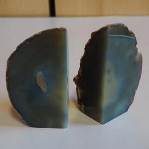 May include: Two agate bookends. Each bookend is a half-circle shape with a flat edge. The colours are shades of blue, green, and brown, with a natural, rough edge. The bookends have a polished face.