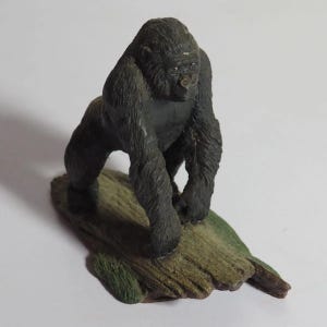 Pode incluir: Uma estatueta de gorila cinza-escuro e preto sobre uma base texturizada em forma de tronco castanho e verde. O gorila é representado numa postura inclinada para a frente, com uma textura de pelo detalhada e uma constituição musculada.