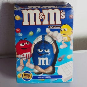 May include: A blue M&M's branded PC mouse packaged in a rectangular cardboard box. The box features graphics of red, yellow, and blue M&M characters, the text "m&m's" and "PC Mouse," and a small label indicating 125 g M&M's.