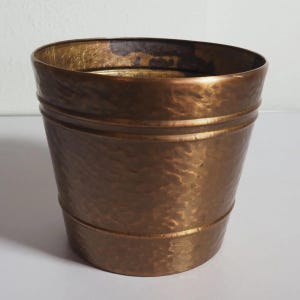 Puede incluir: Una maceta de metal redonda y cónica con textura martillada y un acabado en bronce bruñido cálido. El recipiente presenta dos bandas horizontales en relieve que rodean el exterior, con un borde interior liso y oscuro en la parte superior.