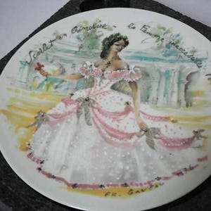 Könnte beinhalten: Dekorative Porzellanteller mit handgemalter Illustration einer Frau in einem rosa-weißen Kleid. Der Teller trägt den Text "Scarlet et Guimoline - la Femme inaccessible 1865". Ein Vintage-Sammlerstück.