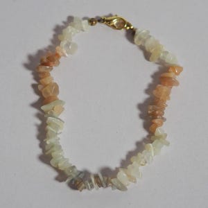 May include: A bracelet composed of irregular, tumbled stone chips in shades of pale peach, cream, and translucent white. The stones are strung together in a circular arrangement and secured with a gold-toned metal lobster claw clasp at the top.