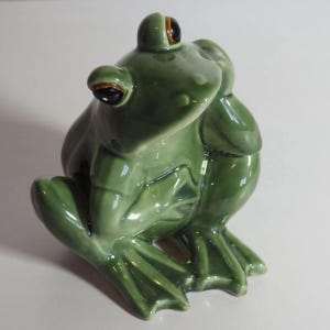 Könnte beinhalten: Eine glänzende, salbeigrüne Froschfigur aus Keramik in sitzender Position. Der Frosch hat große, dunkle, runde Augen mit braunen Akzenten, eine glatte Oberfläche und detaillierte Schwimmfüße. Er ist in einer nachdenklichen Pose dargestellt.