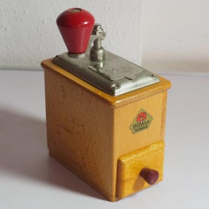 May include: A wooden coffee grinder with a light brown finish, featuring a red handle knob and a small red knob on the front drawer. The top has a silver metal plate with a crank mechanism. A diamond-shaped label on the front reads Dienes Mokka.