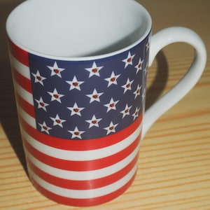 Può includere: Tazza in ceramica bianca con il design della bandiera americana. La tazza ha un manico e un interno bianchi. Il design esterno comprende strisce rosse e bianche e un campo blu con stelle bianche, ciascuna con un centro rosso. Una tazza a tema patriottico.
