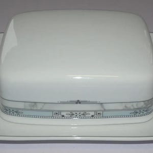 Può includere: Burriera in ceramica bianca con coperchio e base quadrati. Il piatto presenta una fascia decorativa con un motivo marmorizzato azzurro e grigio. I bordi del piatto sono delineati da una sottile linea nera.