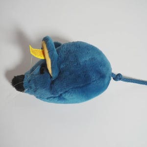 Op de afbeelding: Een blauwe pluche muis met een zwarte neus en snorharen. De muis heeft gele accenten op de oren en een blauwe touwstaart. Een geel label is aan het oor bevestigd.