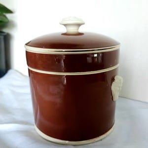 Op de afbeelding: Een bruine keramische pot met een deksel en een witte knop. De pot heeft een cilindrische vorm met een witte rand aan de basis en bovenkant. Een wit decoratief element bevindt zich aan de zijkant. De pot wordt waarschijnlijk gebruikt voor opslag.