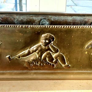 May include: A rectangular brass planter with embossed cherub figures. The planter features a central cherub figure reclining and holding a tool, flanked by two cherubs with wings. The planter is placed on a light-coloured surface.