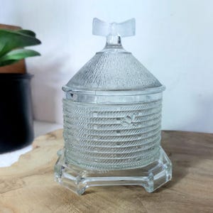 Op de afbeelding: Een doorzichtige glazen honingpot in de vorm van een bijenkorf met een textuur van ribbels aan de buitenkant en een afneembaar deksel. Het deksel heeft een kleine, matglazen handgreep in de vorm van een strik. De voet is zeshoekig.
