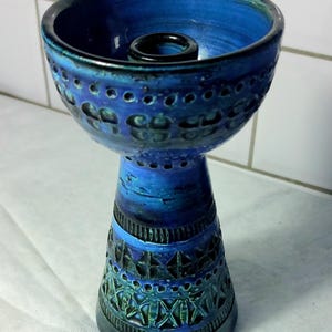 Może przedstawiać: Niebieski ceramiczny świecznik z szeroką misą i wąską, wzorzystą podstawą. Misa ma ciemne wnętrze i jest otoczona teksturowaną powierzchnią z małymi otworami. Podstawa ma geometryczne wzory w odcieniach niebieskiego i zielonego.