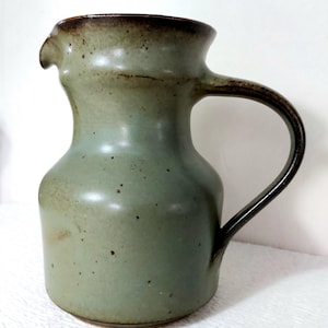 Puede incluir: Una jarra de cerámica con esmalte verde salvia y motas de color marrón oscuro. El recipiente presenta una base ancha y redondeada, un cuello estrecho, un asa curva en el lado derecho y un pequeño pico vertedor en el borde superior marrón.