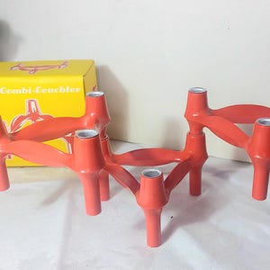 Op de afbeelding: Twee rode kandelaars met zilverkleurige inzetstukken. De kandelaars hebben een uniek, geometrisch ontwerp met meerdere armen en poten. Een gele doos met de tekst "Combi-Leuchter" staat op de achtergrond.
